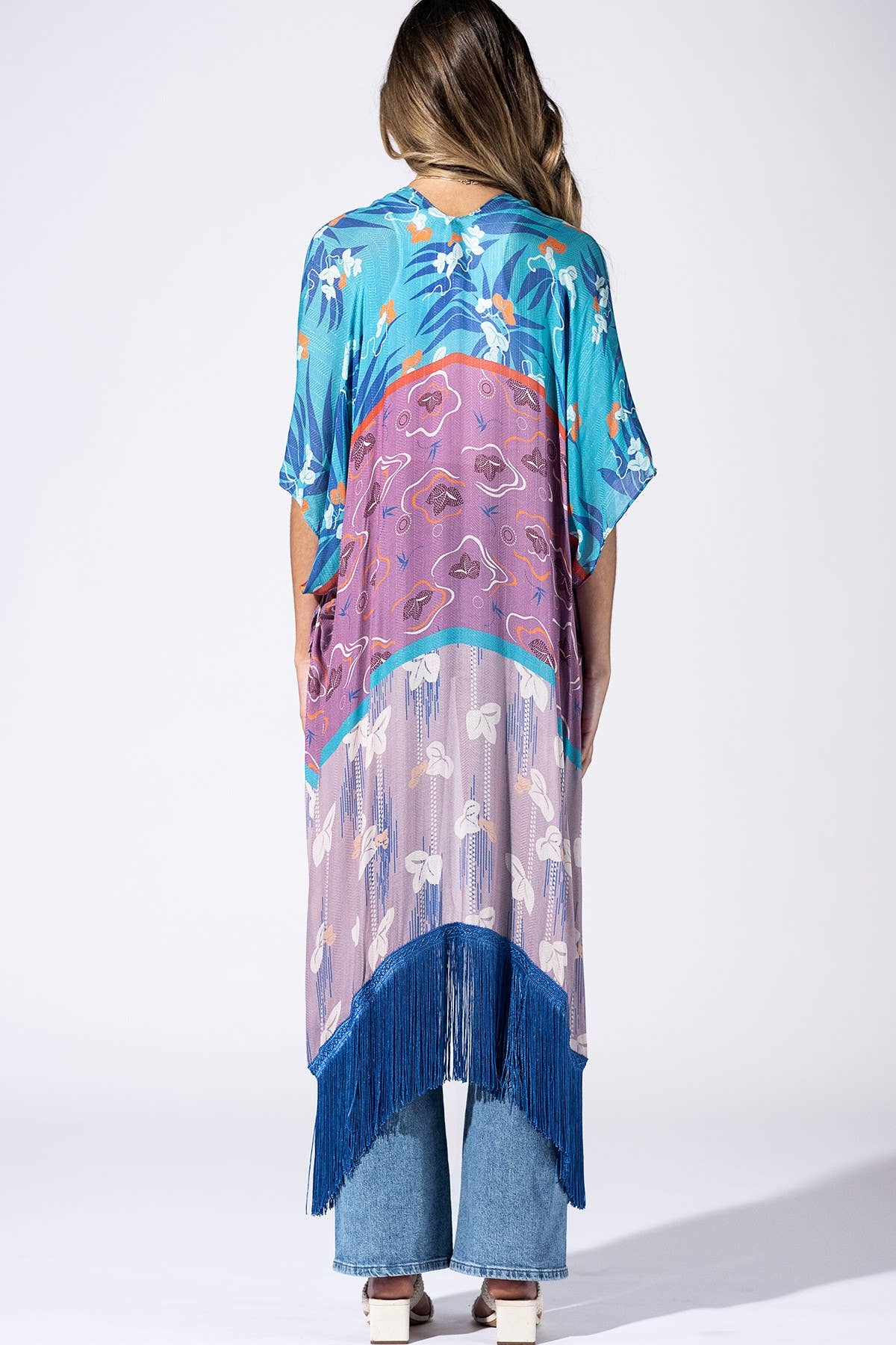 A woman viewed from behind wearing a loose, short-sleeve kaftan-style cover-up with a multicolor tropical and floral print, blue fringe along the hem, and light blue jeans, photographed against a plain white studio background.
