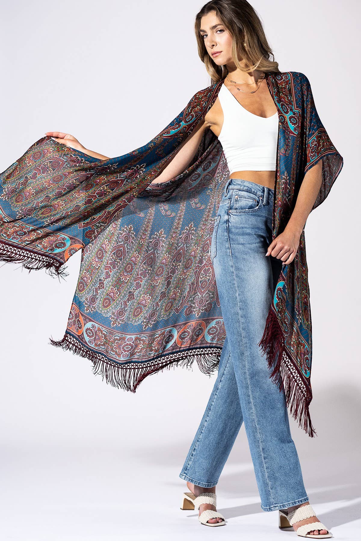 A woman modeling a lightweight, multicolored paisley shawl with fringe, worn open over a white crop top and blue jeans, against a plain white studio background.