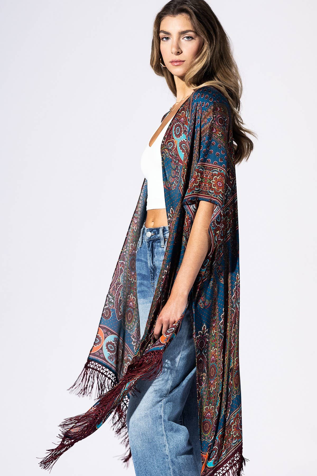 A woman modeling a long, open-front paisley shawl or kimono in deep blue, red, and orange tones with fringed edges, worn over a white crop top and light blue jeans against a plain white studio background.