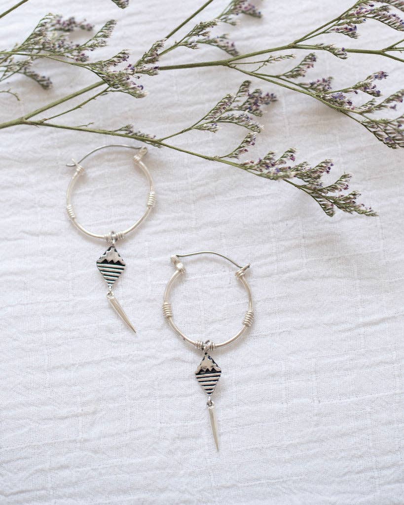Silver hoop earrings with geometric designs on a white fabric background with dried flowers.