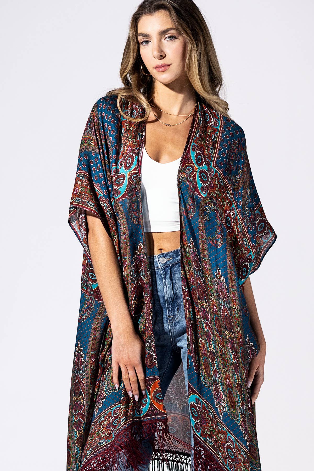 A woman modeling a long, open-front bohemian-style kimono cover-up with a complex paisley and floral pattern in teal, rust red, brown, and black, worn over a white crop top and blue jeans against a plain white studio background.