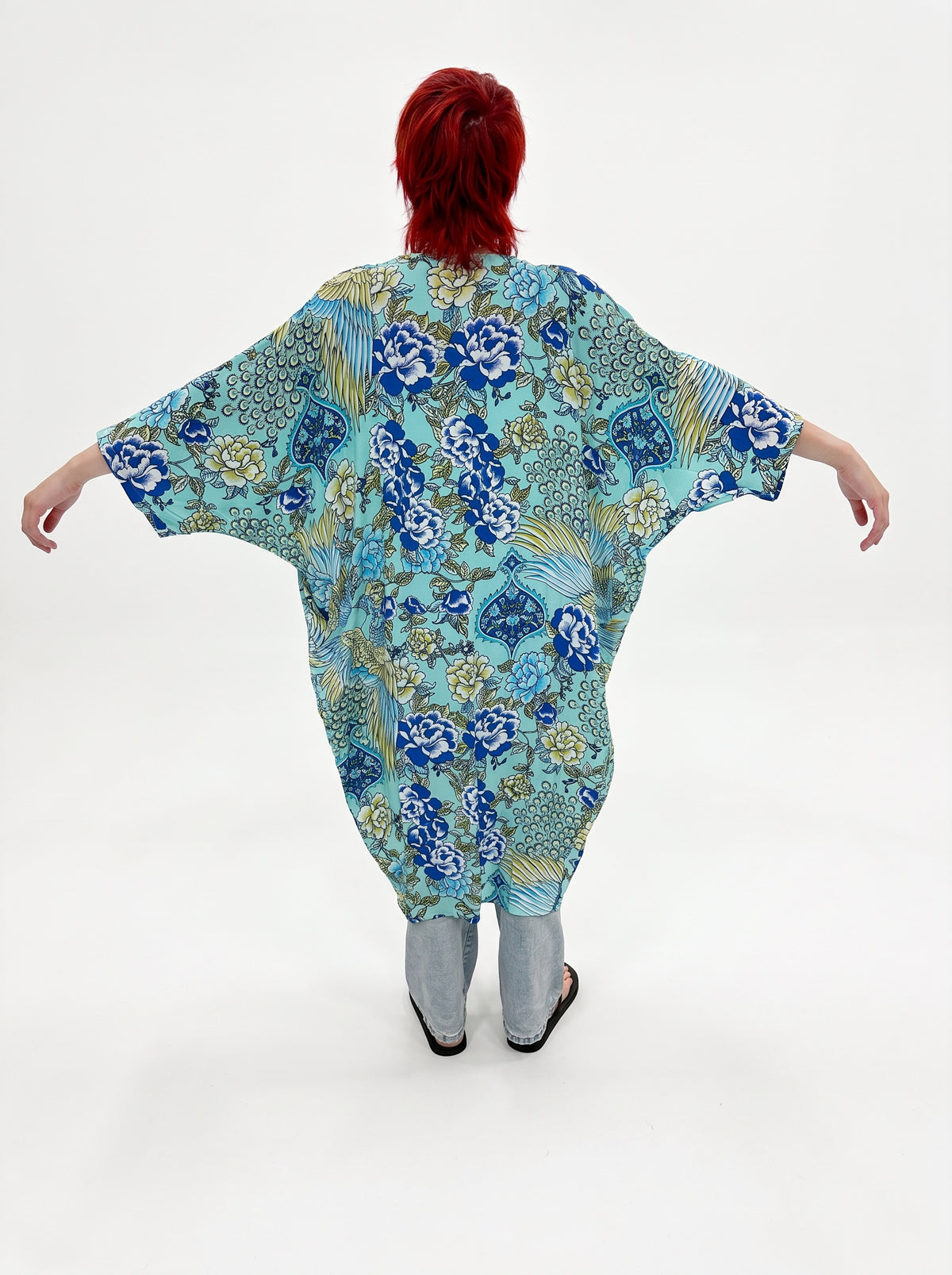 A person with bright red hair stands with arms outstretched, viewed from the back, wearing a loose, kaftan-style top with a blue, teal, and gold floral and ornamental print. The top is paired with light gray pants and black sandals against a plain white studio background.