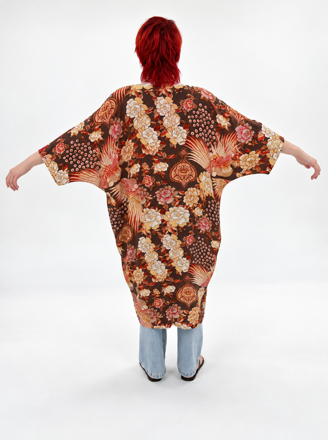 A person viewed from behind wearing a loose, short-sleeve kaftan-style top with a dense floral and botanical print in brown, cream, pink, and orange tones, paired with light blue jeans against a plain white studio background.