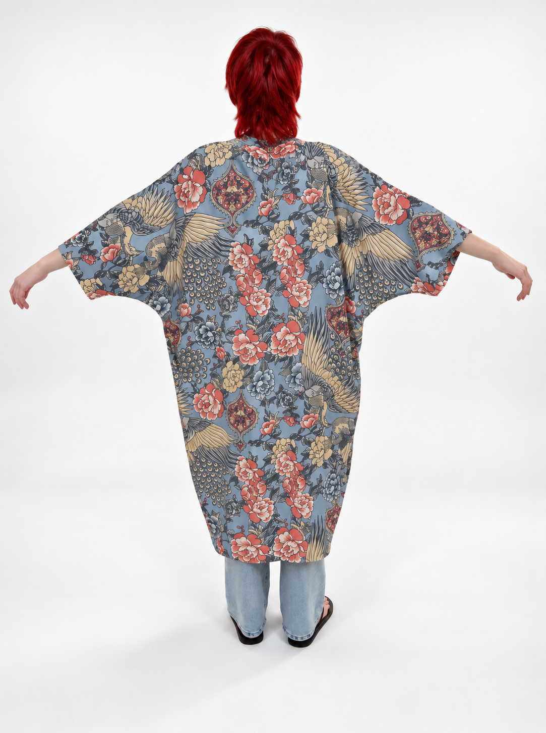 A person with bright red hair stands with arms outstretched, viewed from the back, wearing a loose, oversized kaftan-style top with a blue-gray base and an allover floral and bird pattern in red, beige, and gold. The person is also wearing light blue jeans and black shoes against a plain white studio background.