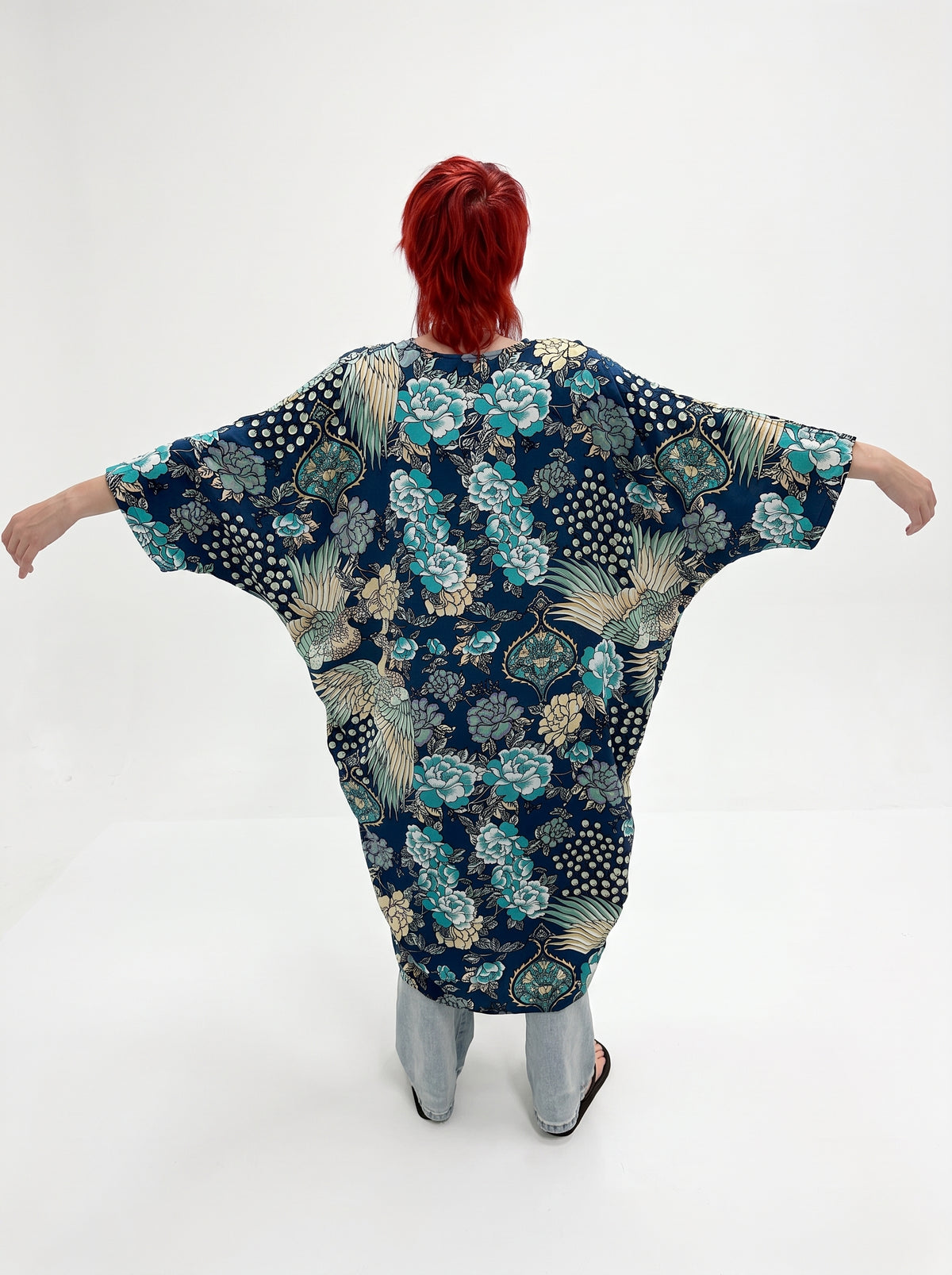 A person with bright red hair stands with arms outstretched, viewed from behind, wearing a loose, cape-like patterned top. The garment has a dark navy base with teal, beige, and white floral and leaf motifs.
