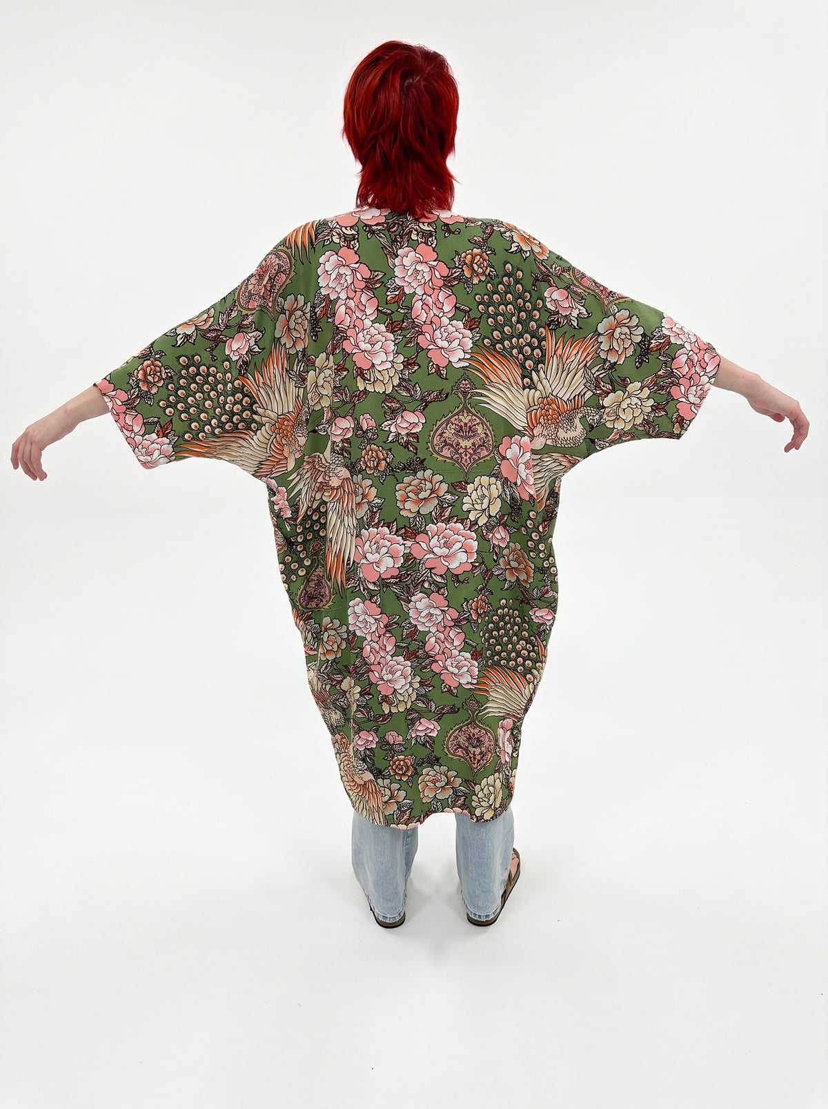 A person viewed from behind wearing a loose, short-sleeve kaftan-style top with a dense floral and botanical print in green, cream, pink, and orange tones, paired with light blue jeans against a plain white studio background.