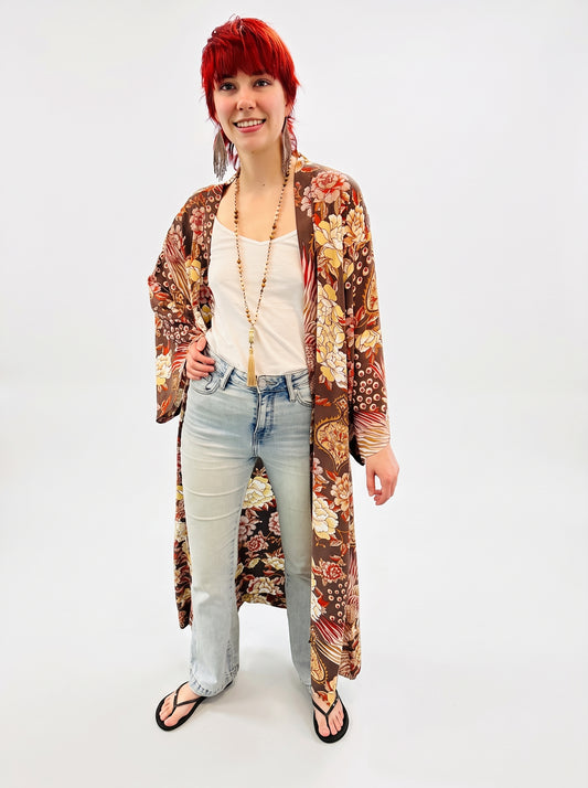 A model with bright red hair stands against a white studio background wearing a long-sleeved, open-front floral kimono in brown, beige, and orange tones over a white tank top, light-wash jeans, and black sandals.