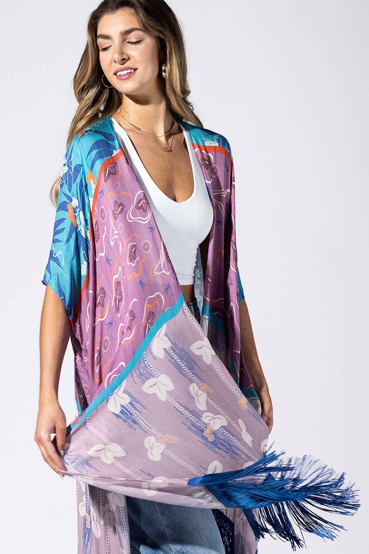 A woman models a lightweight, open-front kimono-style cover-up with a vibrant multicolor tropical print and blue fringe, worn over a white tank top and blue jeans against a light gray studio background.