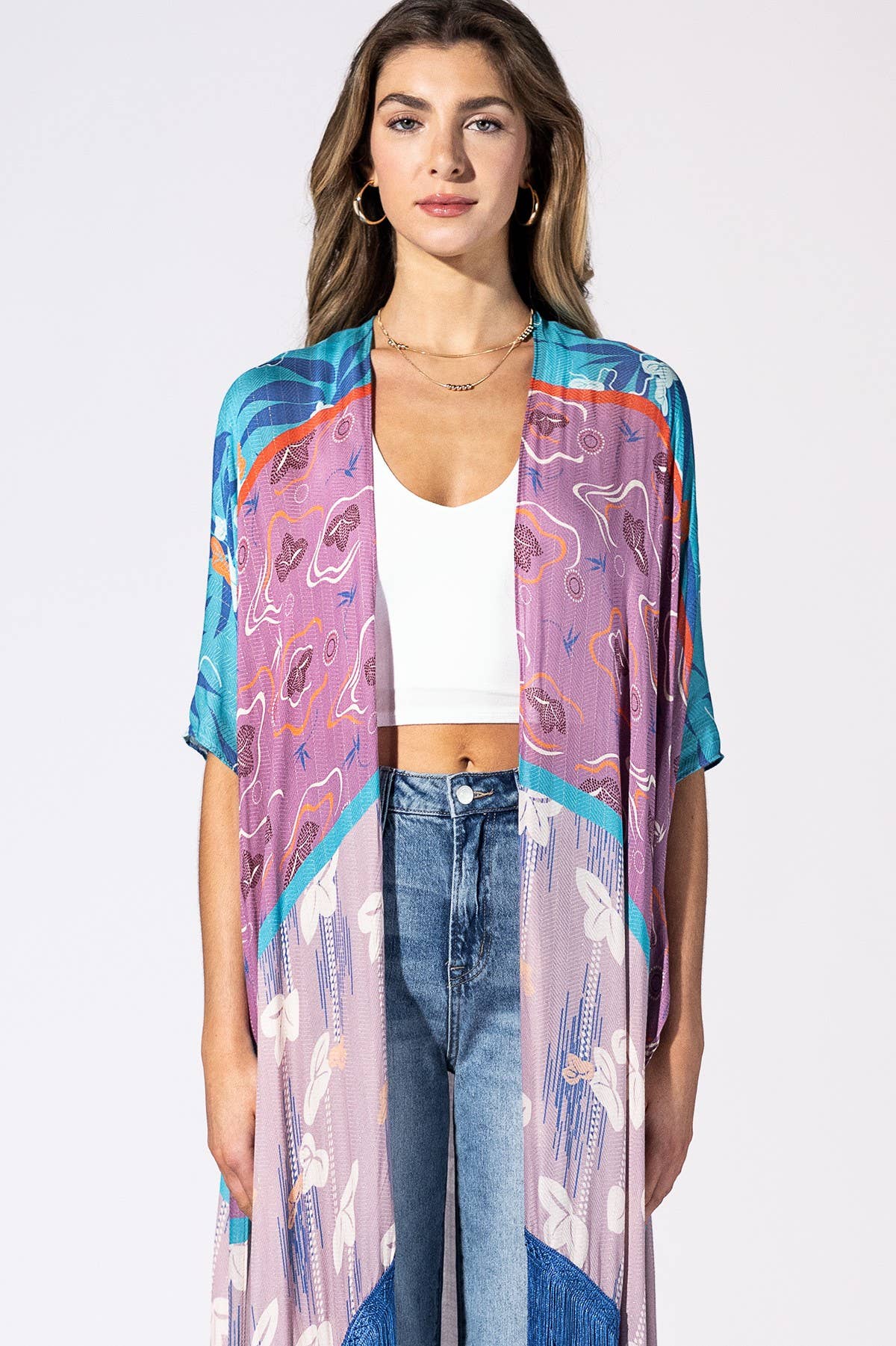 A woman modeling a lightweight open-front kimono cardigan with a vibrant abstract print in shades of purple, blue, orange, and white, worn over a white crop top and blue jeans against a plain white studio background.