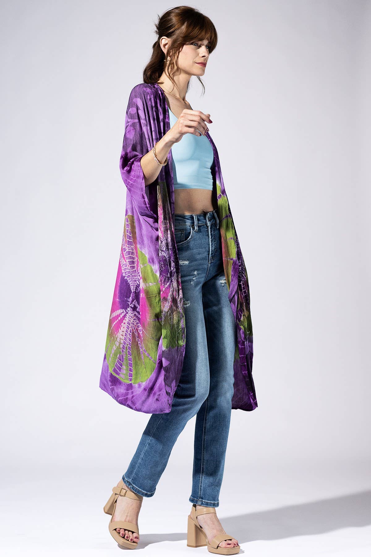 Woman wearing a long, colorful kimono-style garment over a light blue top and jeans on a white background