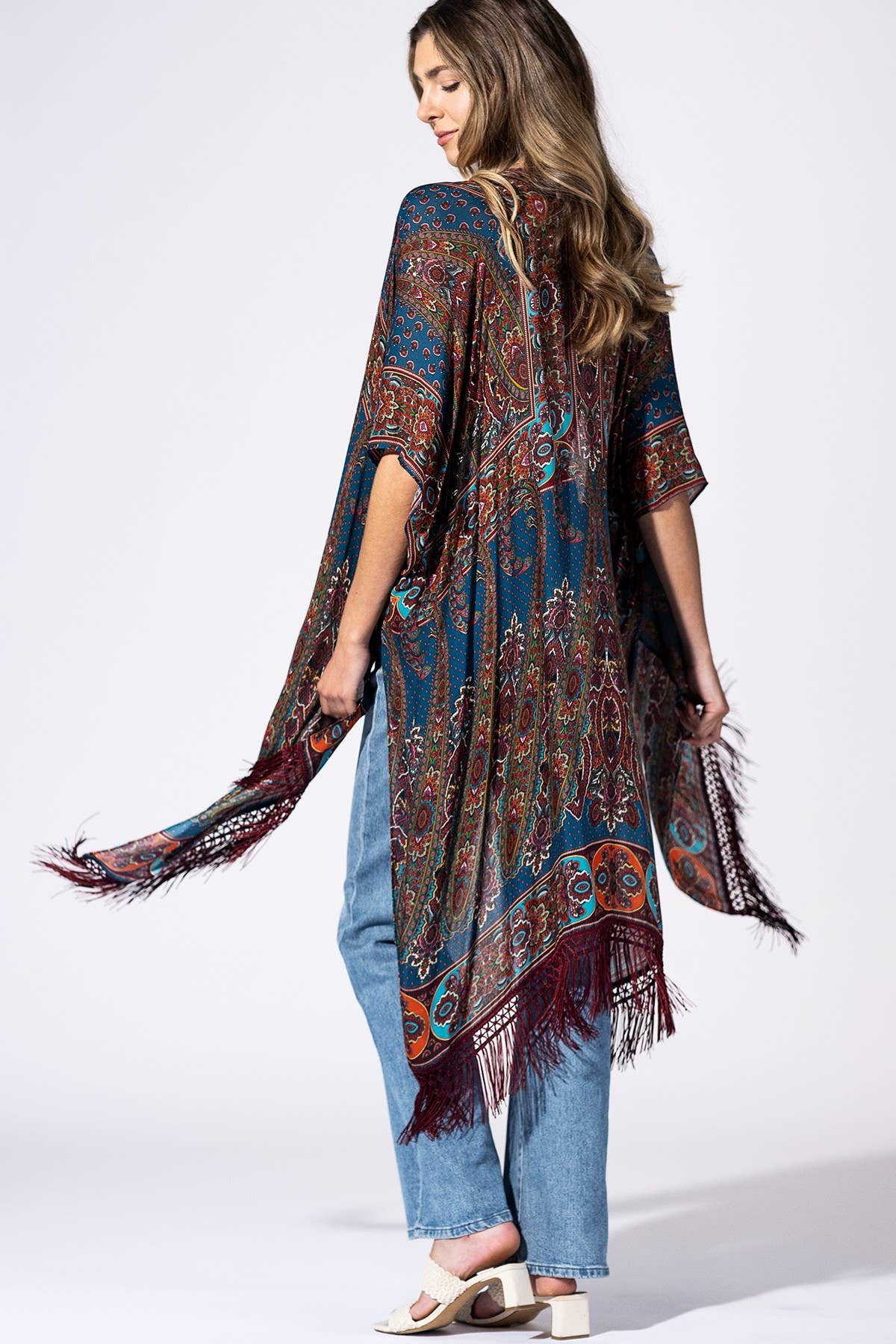 A woman shown from the back wearing a loose, patterned fringed poncho or kimono in blue, burgundy, and orange tones over light blue jeans and white heeled sandals, photographed against a plain light gray studio background.
