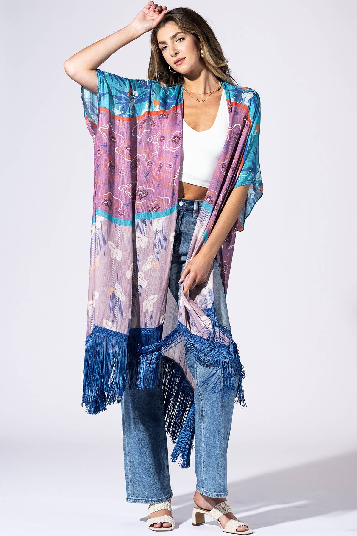 A woman models a lightweight, semi-sheer kaftan-style cover-up with a colorful abstract print in shades of blue, purple, pink, and teal, featuring a long fringe hem. She is styled with a white crop top, blue jeans, and white heeled sandals against a plain white studio background.