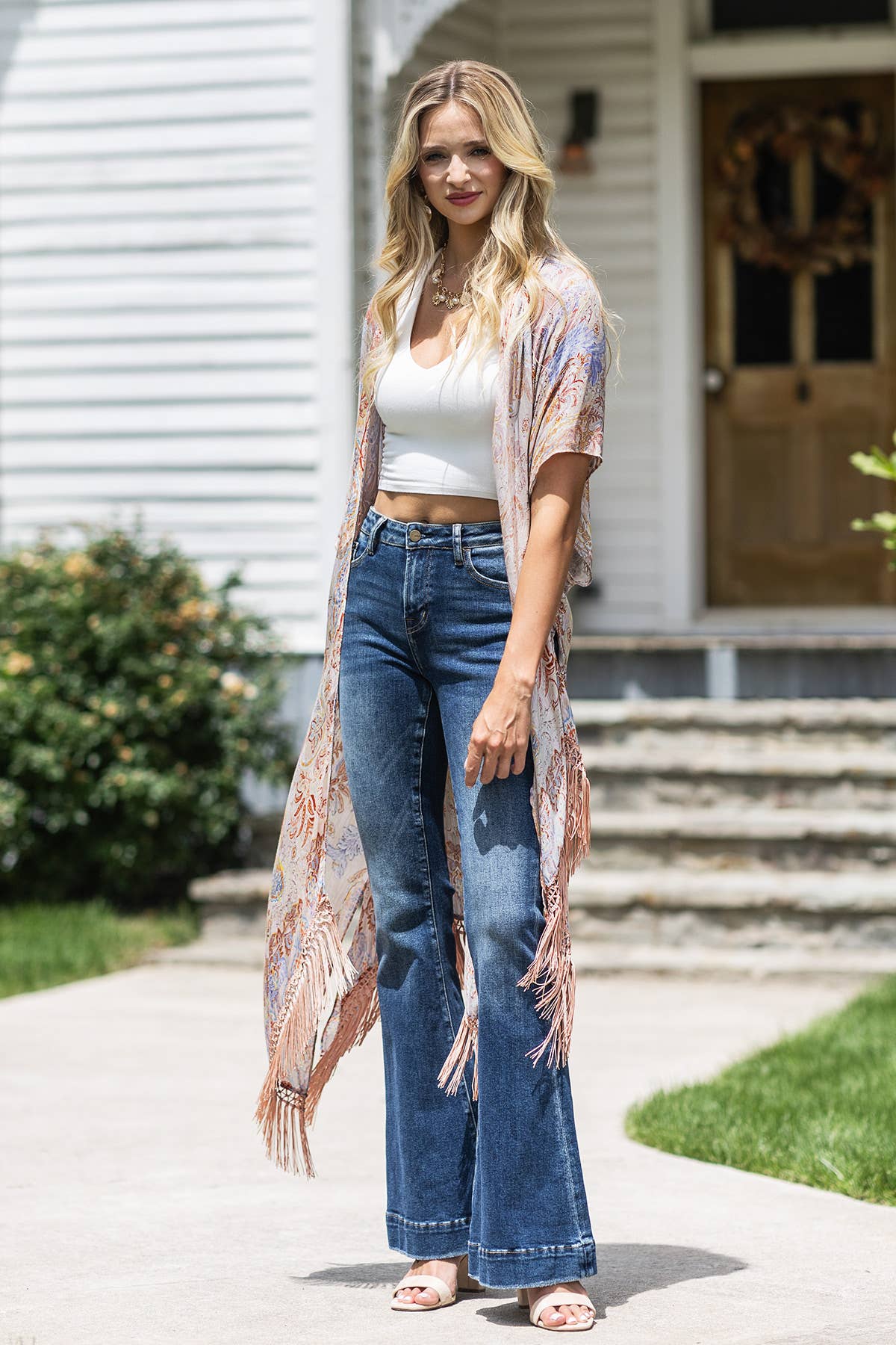 Woman wearing a white top, blue jeans, and a beige kimono-style cardigan standing outside a house.