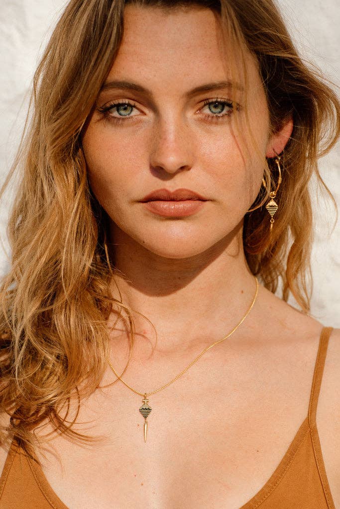 Woman with blonde hair and earrings wearing a brown top against a neutral background