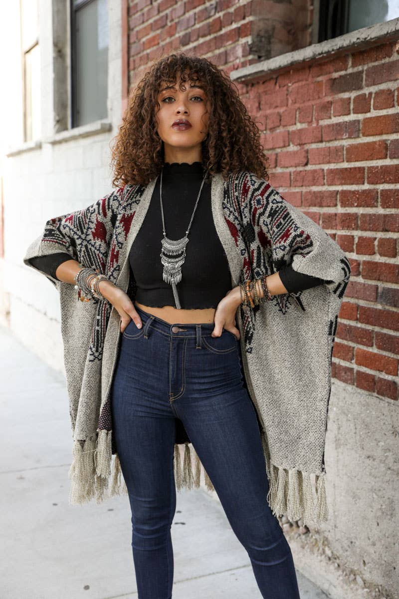 Woman wearing a patterned cardigan, black top, and blue jeans standing against a brick wall.