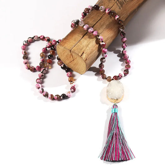 A beaded necklace with pink, brown, and white marbled beads, featuring a large white crystal dome and a colorful tassel with pink, blue, and purple threads, displayed on a wooden block against a white background.