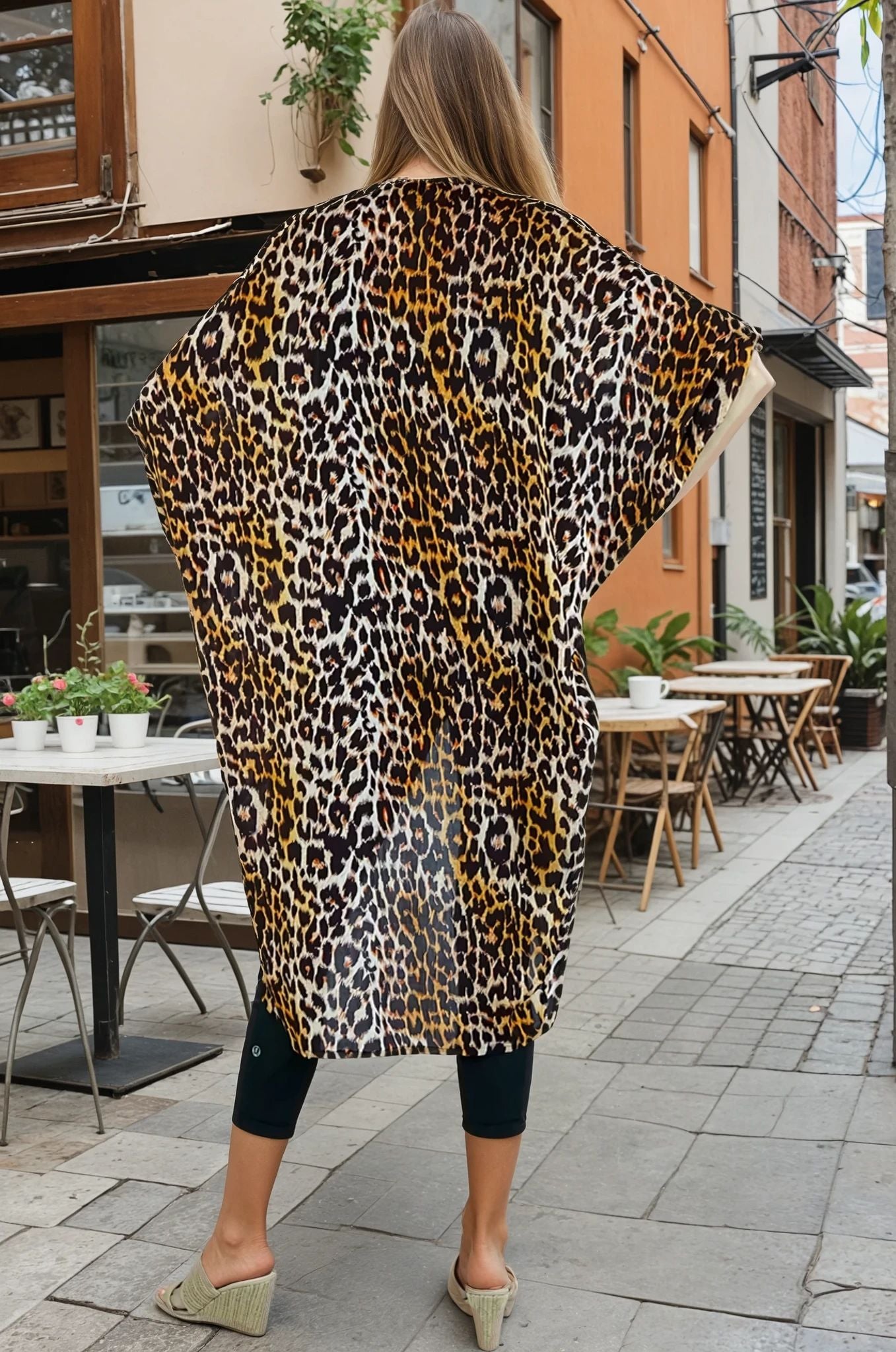 Person wearing a leopard print kimono walking on a street with tables and chairs.