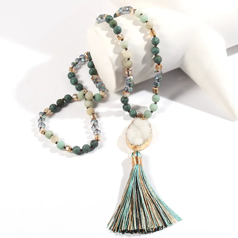 A bohemian-style beaded necklace with green, gray, and beige stones, a large white stone pendant, and a long striped tassel, displayed on a white jewelry stand.