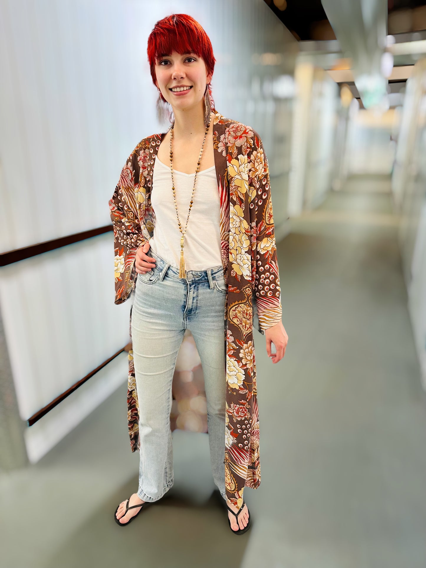 A woman with bright red hair stands in a hallway wearing a long-sleeved, open-front floral kimono cardigan over a white tank top and light-wash jeans.
