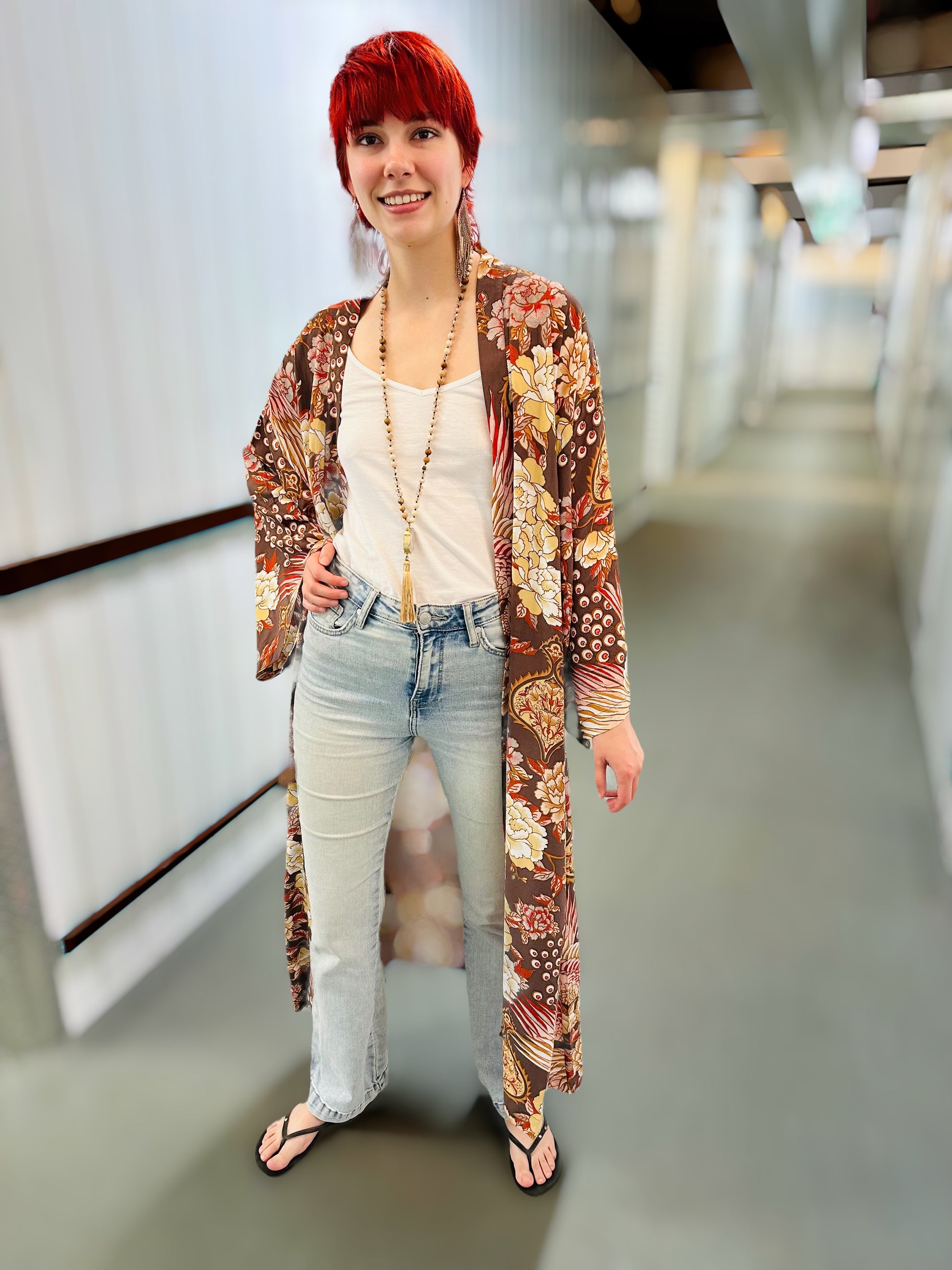 A woman with bright red hair stands in a hallway wearing a long-sleeved, open-front floral kimono cardigan over a white tank top and light-wash jeans.