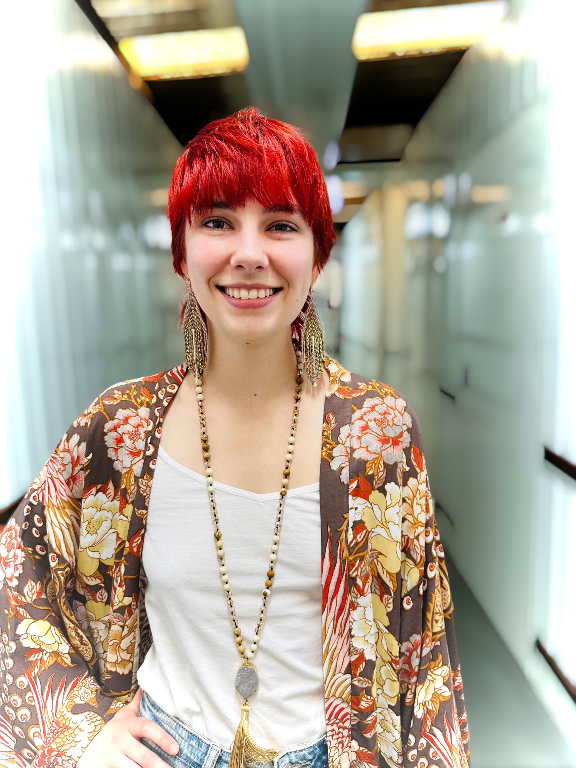 A smiling woman with bright red hair wearing a white tank top, a long beaded necklace, and a brown floral kimono jacket, standing in a brightly lit hallway.