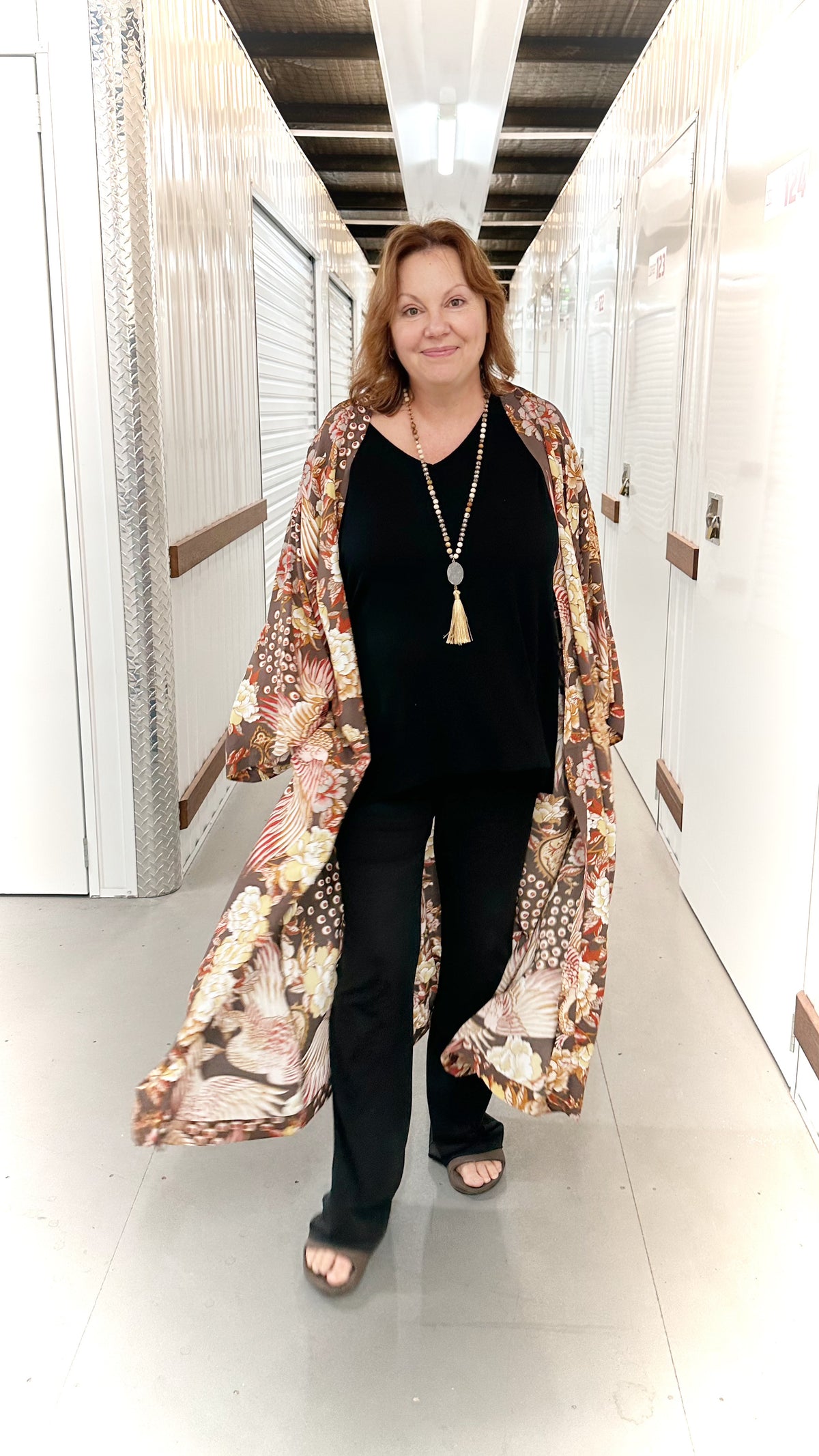 A woman walking down a hallway wearing a long-sleeved, open-front kimono-style coat with a brown, beige, and black floral pattern over a black top and black pants.