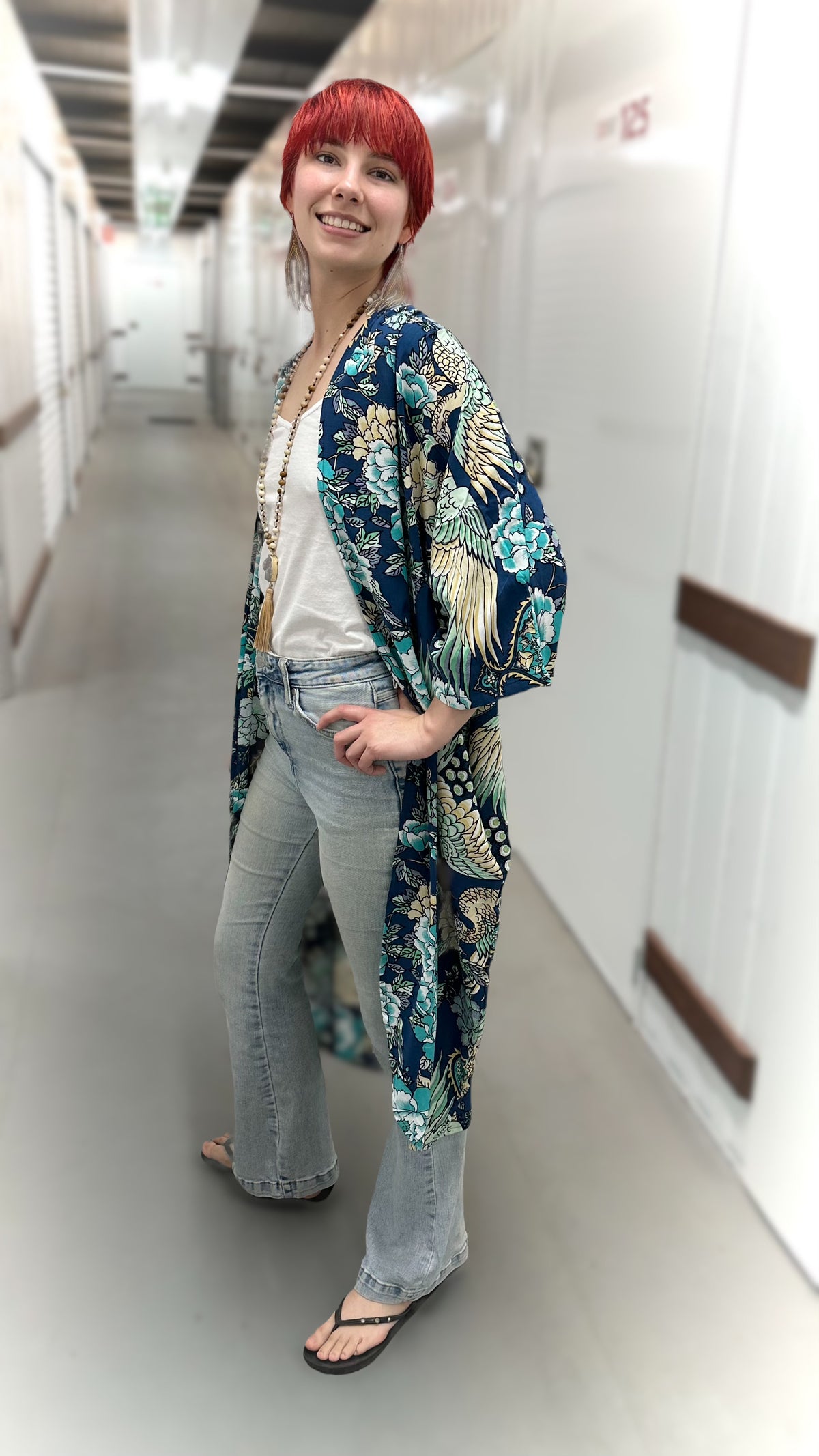 A woman with bright red hair stands in a hallway wearing a long-sleeved open-front kimono-style cardigan with a blue, green, and beige tropical floral print, paired with light-wash jeans and black flip-flops.