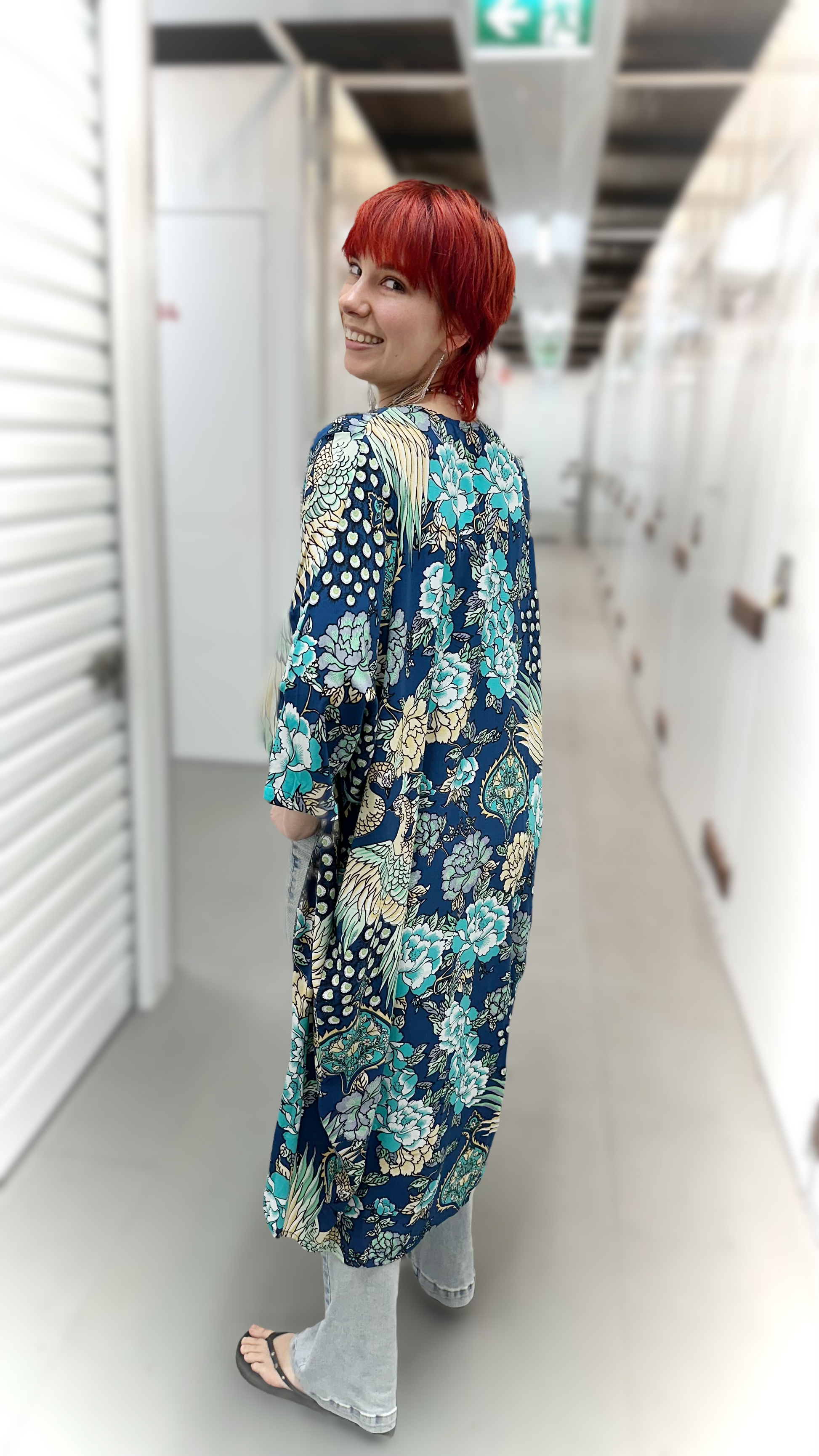 A woman with bright red hair is shown from behind wearing a long-sleeved, knee-length tunic with a dense floral and botanical pattern in navy blue, teal, and beige. She is standing in a hallway with white walls and light-colored flooring.