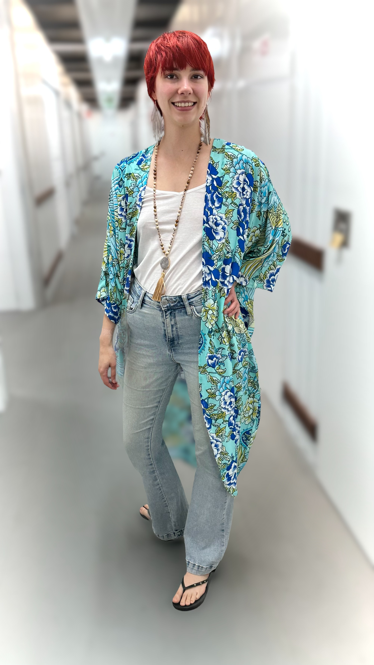 A woman with short red hair walks down a hallway wearing a blue and green floral kimono cardigan over a white tank top, light wash jeans, and black sandals.