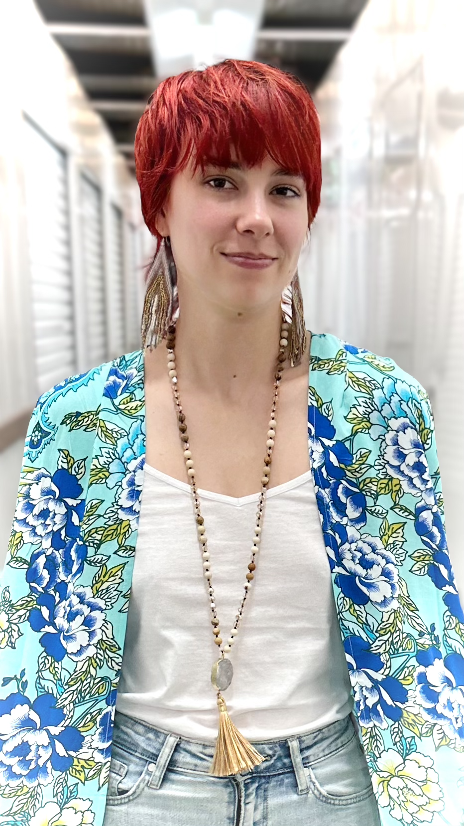 A woman with short red hair wearing a light blue floral cardigan over a white tank top and light-wash jeans, accessorized with a long beaded necklace and large earrings.