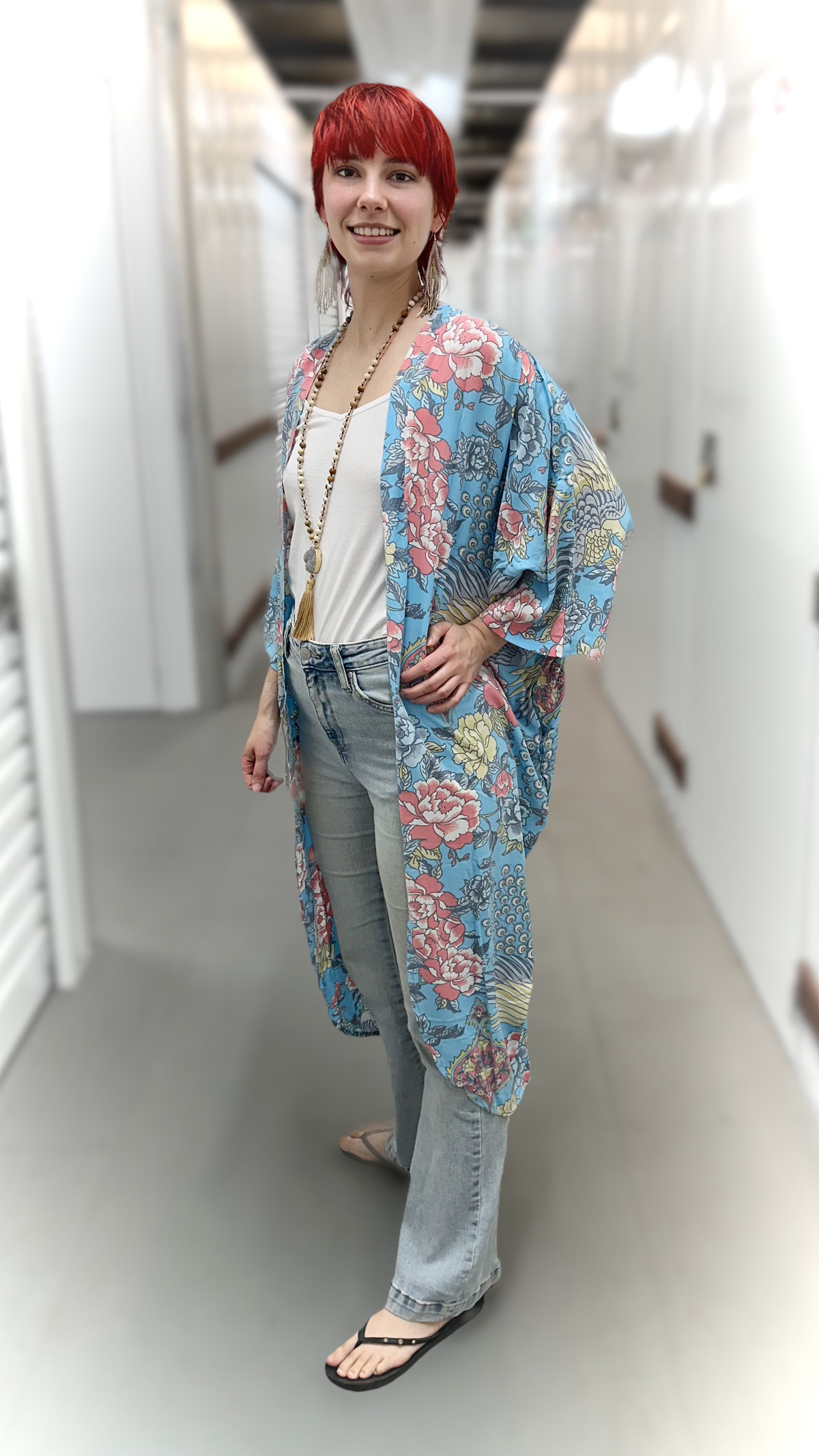 A woman with short red hair stands in a hallway wearing a lightweight open-front kimono-style cardigan with a colorful floral print in blue, pink, and beige over a white top and light-wash jeans.