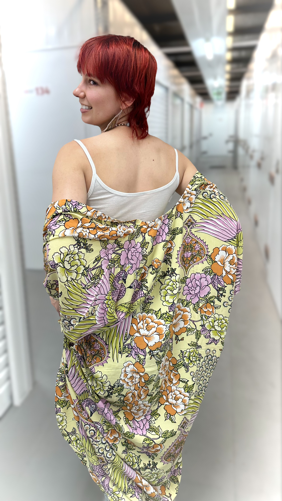 A woman with short red hair is shown from behind, wearing a white camisole and wrapped in a large, draped fabric featuring a colorful floral and botanical pattern on a pale yellow background.