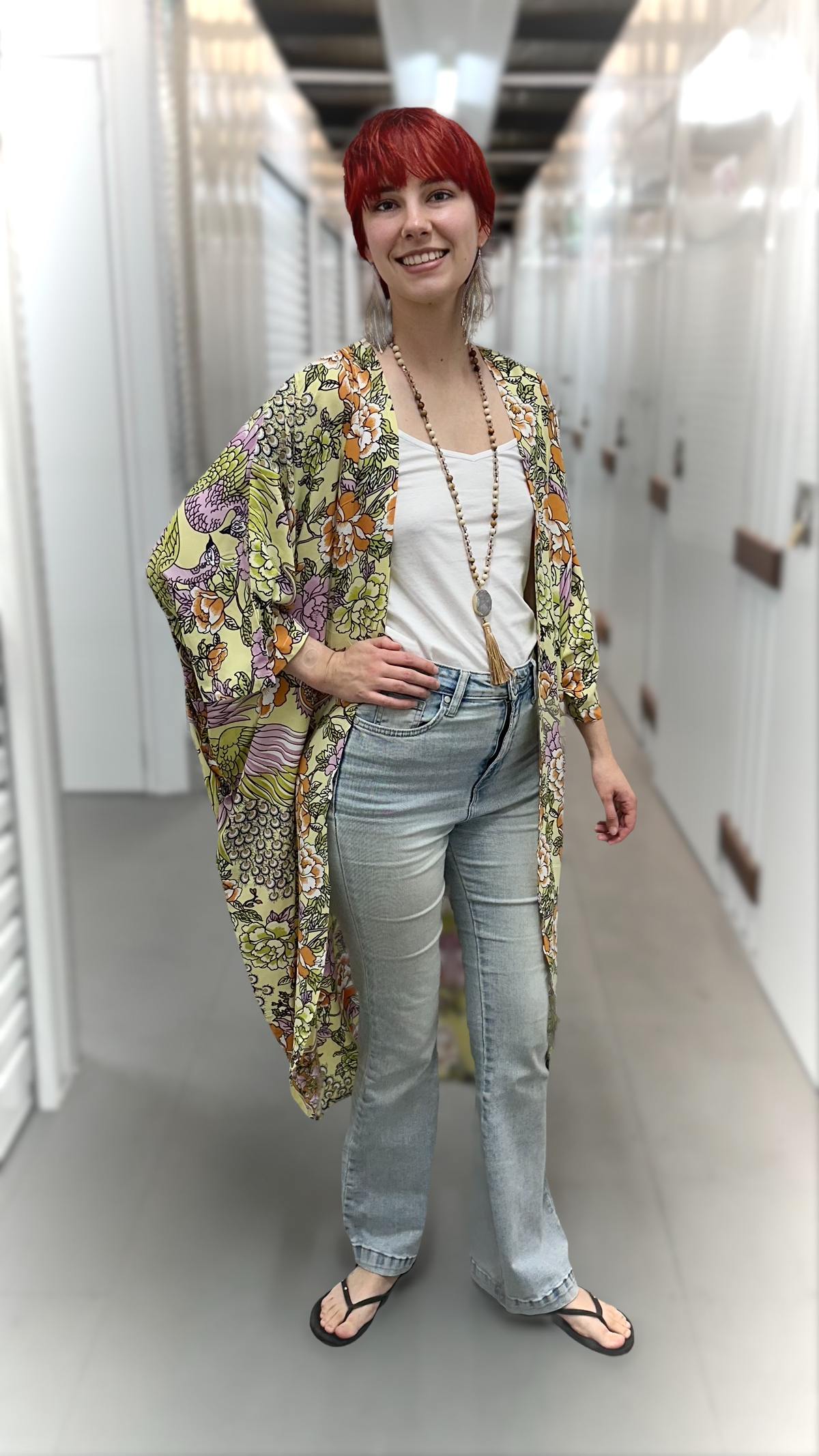 A woman with short red hair stands in a hallway wearing a lightweight open-front kimono-style cardigan with a busy multicolor botanical and floral print in green, yellow, orange, and purple over a white tank top and light blue jeans.