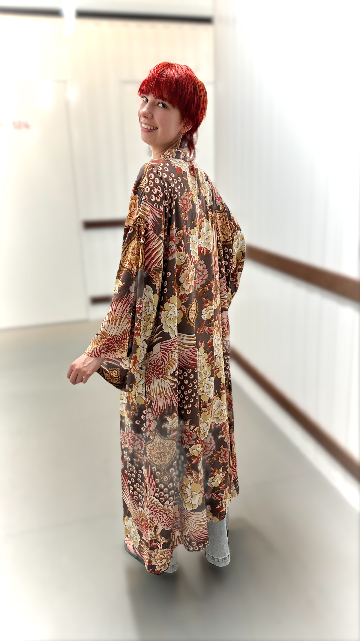 A woman with short red hair is shown from the back wearing a long-sleeved kimono-style robe with a complex multicolor pattern of red, gold, brown, and beige. She is standing in a brightly lit indoor hallway with white walls and a wooden railing.