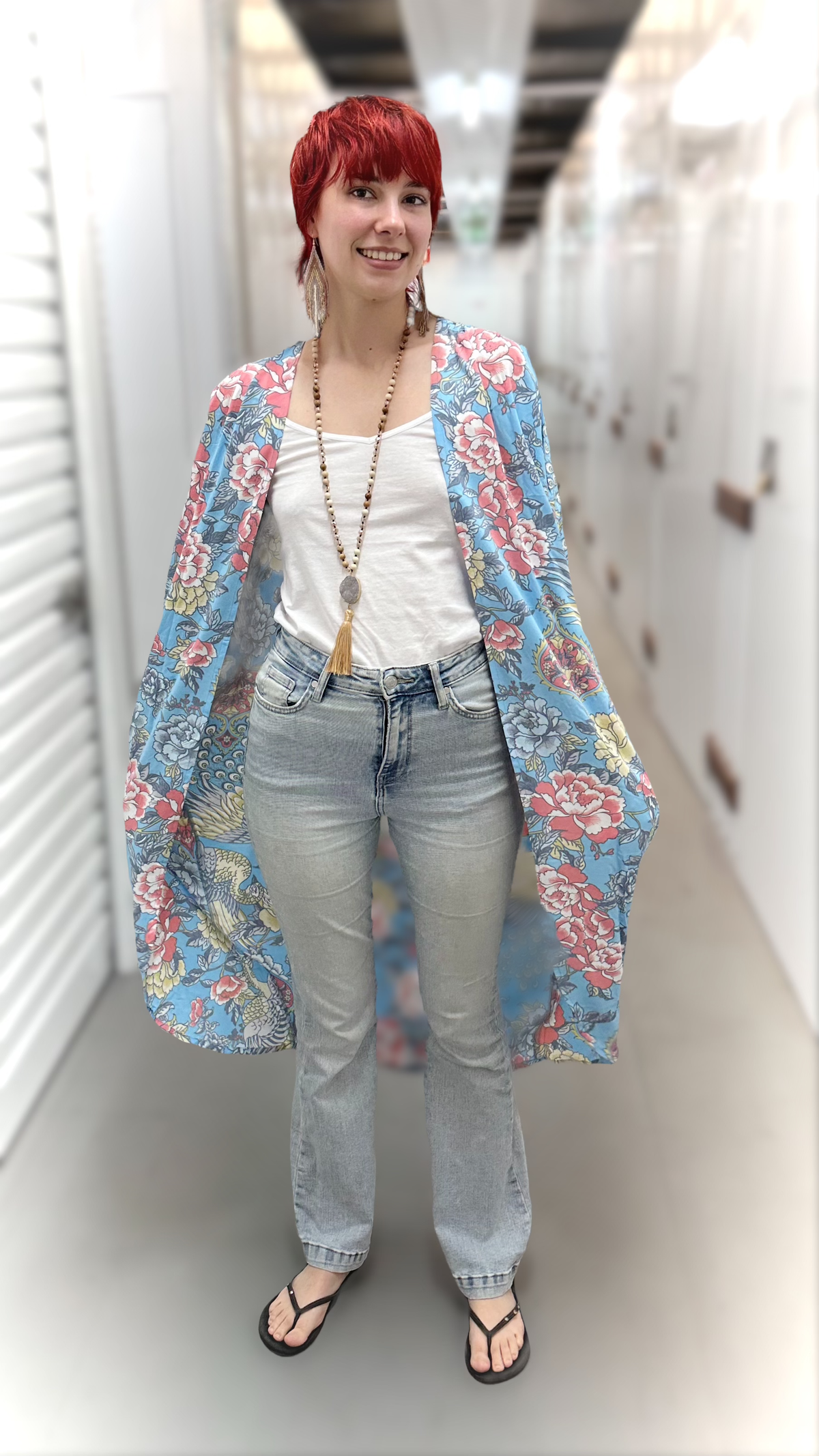 A woman with short red hair stands in a hallway wearing a light blue floral kimono cardigan over a white tank top, light-wash jeans, and black flip-flops.