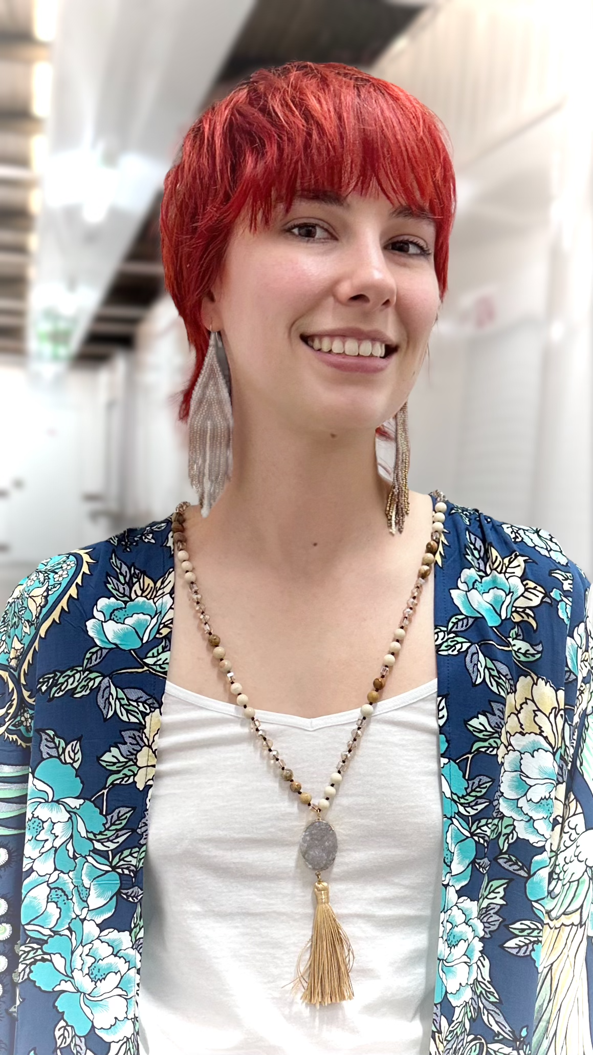 A smiling woman with short bright red hair is shown from the chest up, wearing a white tank top, a blue floral cardigan, and a long beaded necklace with a tassel pendant. She is also wearing long feather earrings.